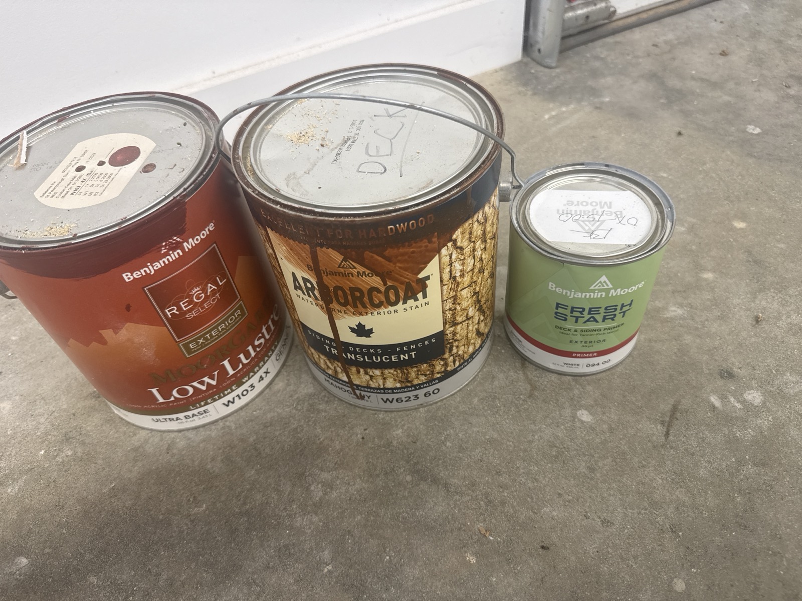Three Benjamin Moore cans on the garage floor: the red exterior paint, the Arborcoat stain marked DECK, and the quart of alkyd primer